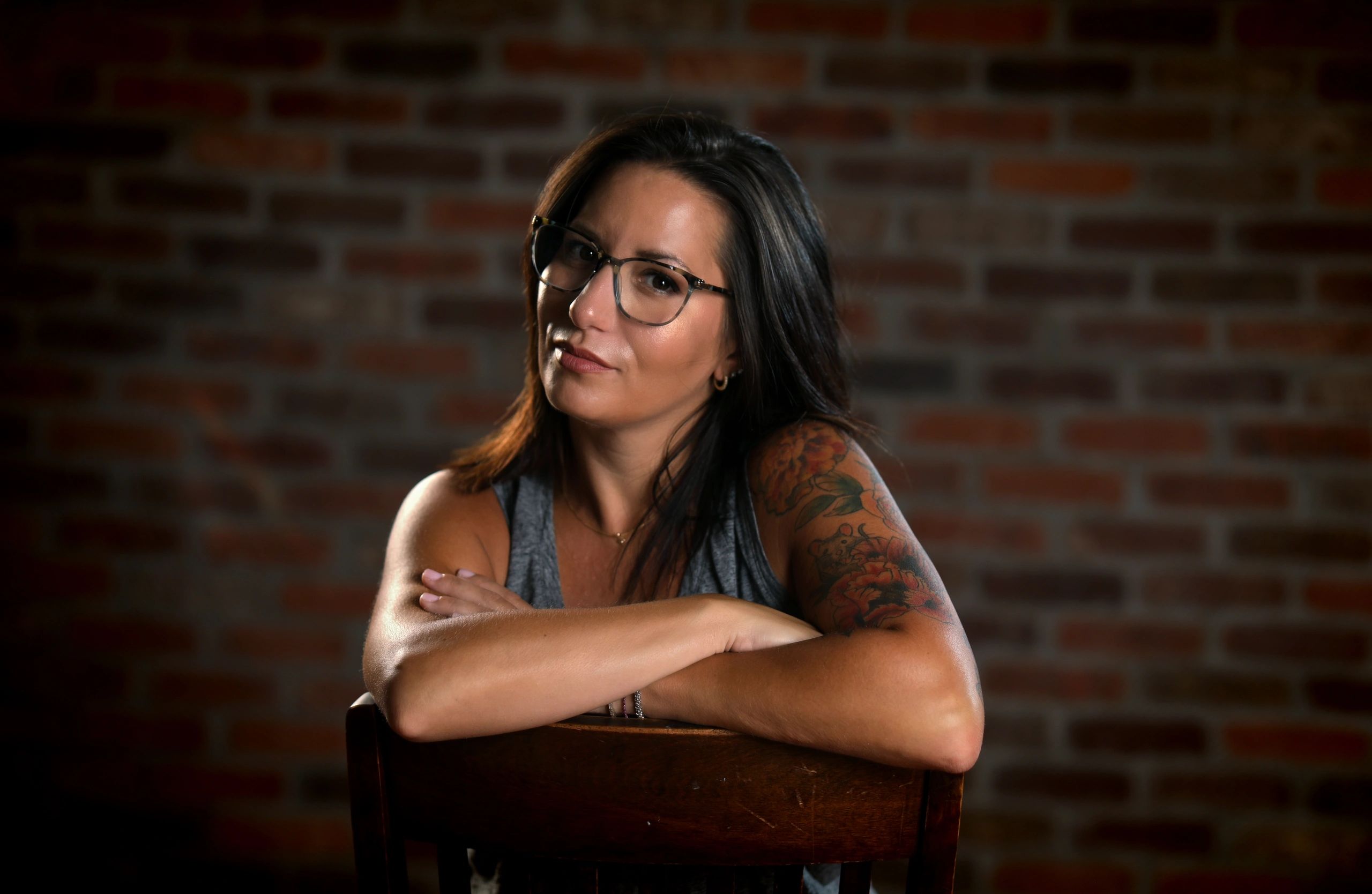 Woman with glasses and tattoo leaning on chair against brick wall.