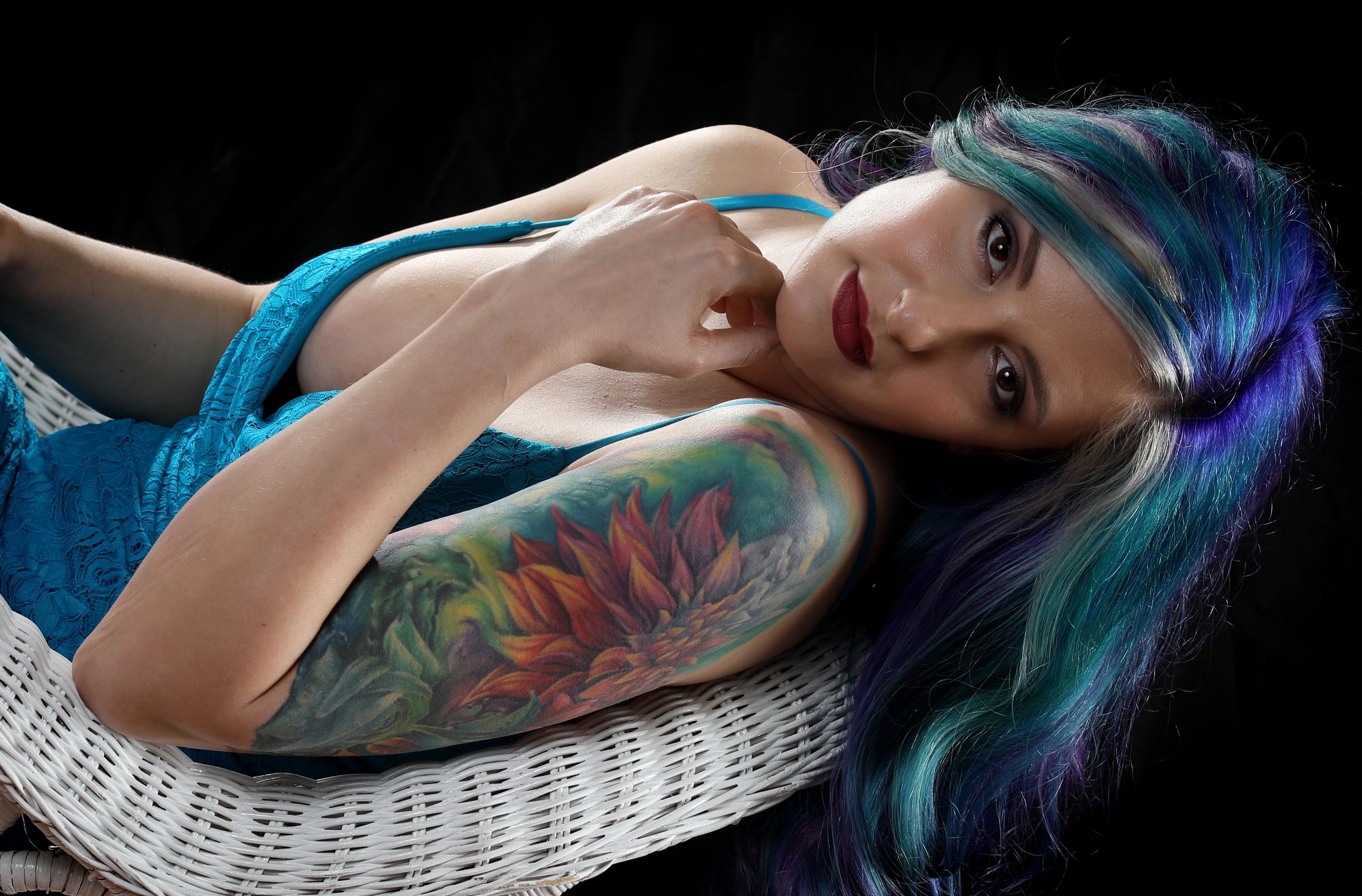 Woman with colorful hair and floral tattoo reclining on white wicker chair.