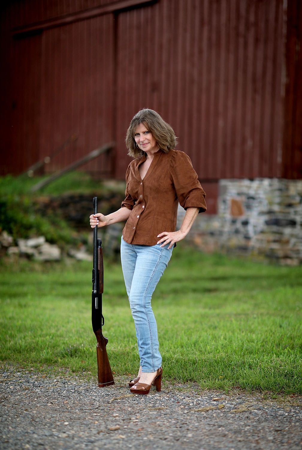 Woman standing outdoors holding a rifle, dressed in jeans and a brown shirt.