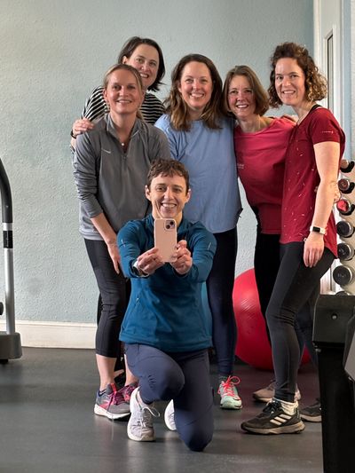 A group of six women smiling and posing for a selfie in a gym.