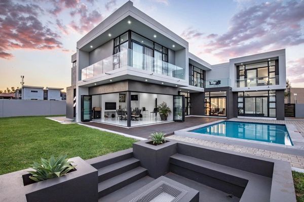 Modern two-story house with glass balconies, a pool, and outdoor seating area at sunset.
