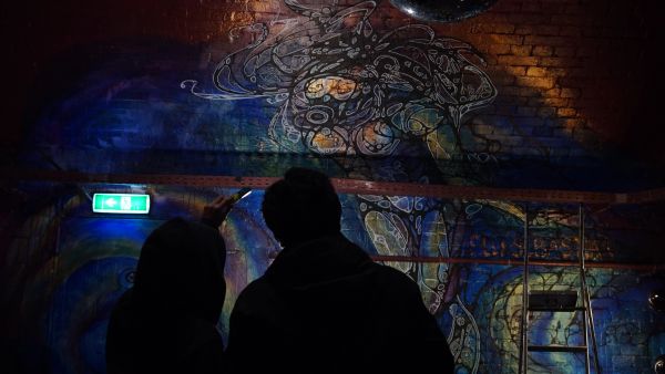 Two people admire a vibrant mural on a dimly lit brick wall.