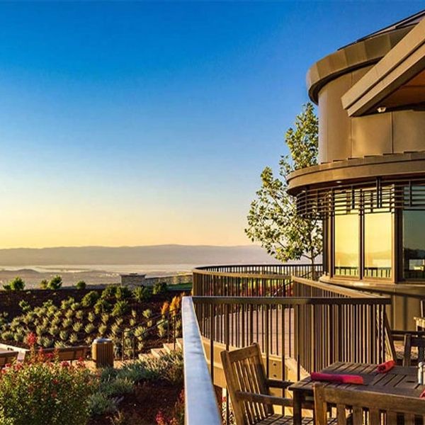 Sunlit balcony with wooden chairs overlooking a scenic vineyard at sunset.
