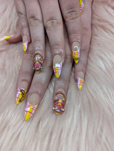 Bright yellow plaid nails with 3D dessert-themed nail art on a soft pink backdrop.
