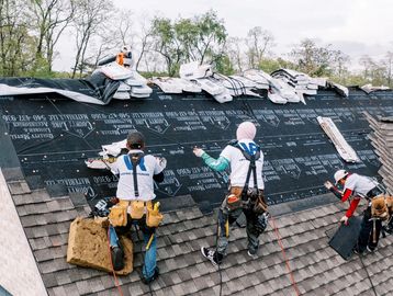 Three workers installing roofing shingles on a sloped roof outdoors.