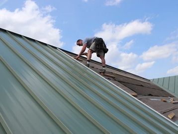 A worker installing green metal roofing under a blue sky.