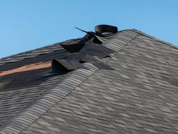 A roof with damaged and lifted shingles under a clear sky.