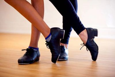 Two dancers wearing black tap shoes crossing their feet on a wooden floor.