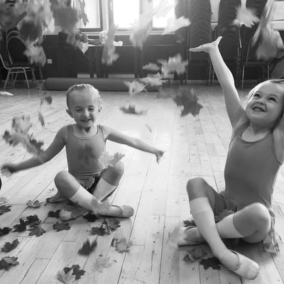 Two young girls joyfully toss autumn leaves indoors while sitting on a wooden floor.