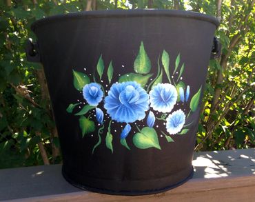 Black bucket with hand-painted blue flowers and green leaves outdoors.