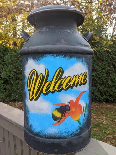 A painted milk can with a 'Welcome' message, bee, and flower.