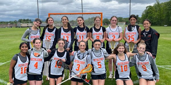 A girls' lacrosse team poses on the field in front of a goal.