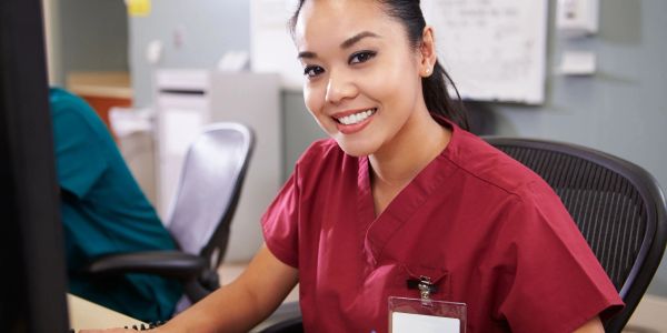 Smiling nurse wearing maroon scrubs sitting at a desk.