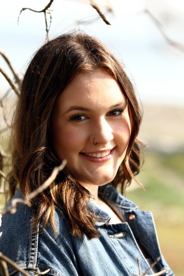 Smiling young woman with brown hair wearing a denim jacket outdoors.