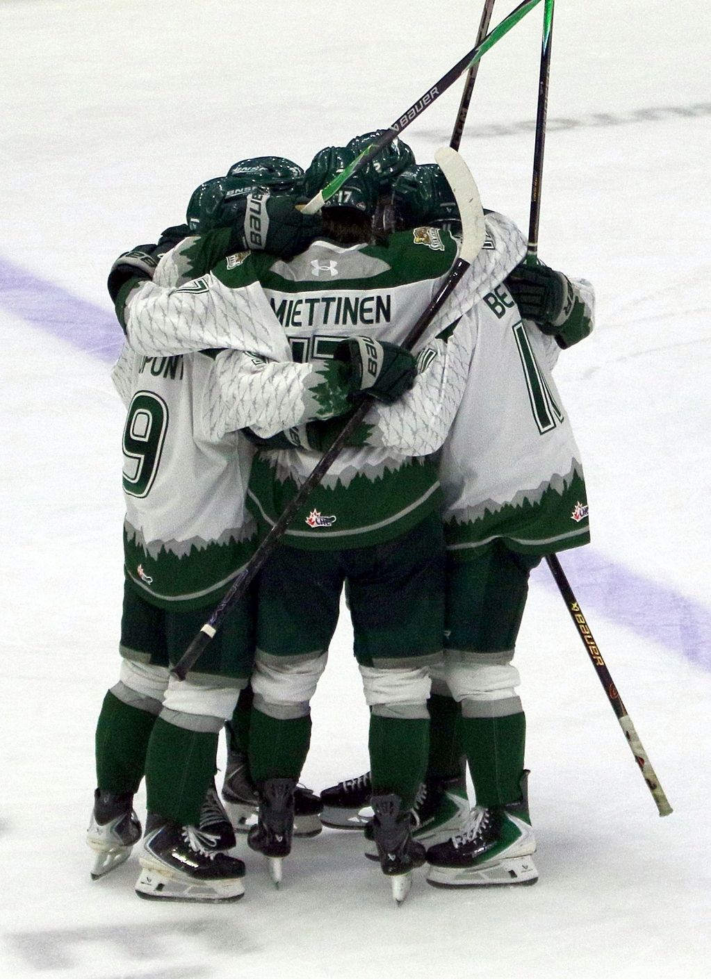 Hockey players in green and white uniforms celebrating with a group hug on the ice.