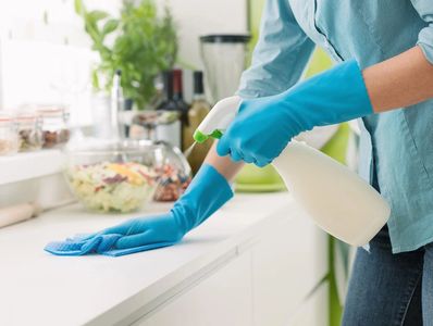 Person cleaning kitchen counter with spray bottle and cloth wearing blue gloves.