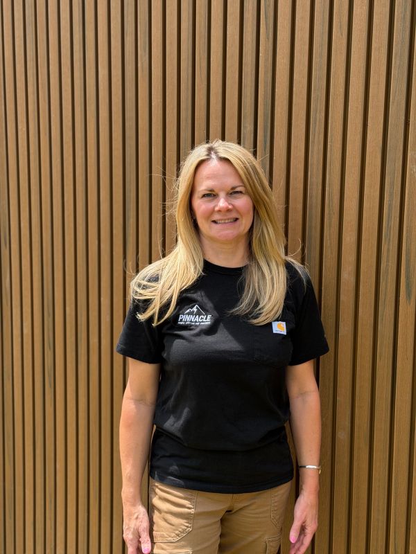 Woman in black 'Pinnacle' shirt standing against wood panel wall.