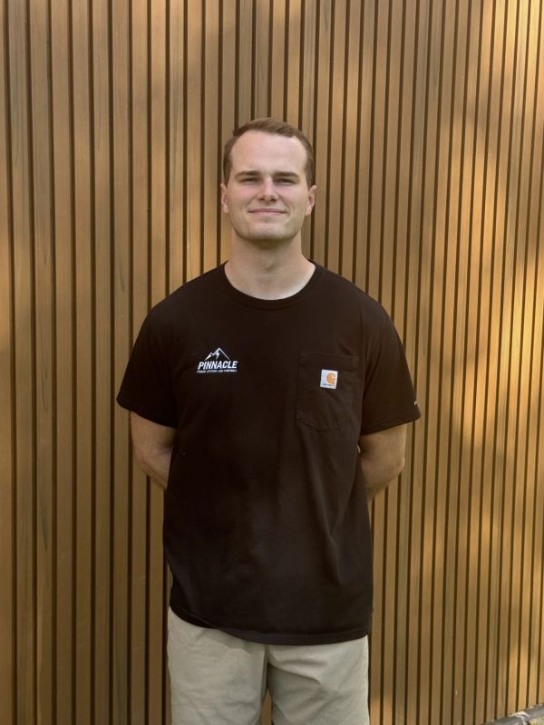 A man stands smiling in front of a wooden slatted wall wearing a black Pinnacle t-shirt.