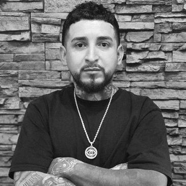 Man with tattoos and necklace poses against stone wall.
