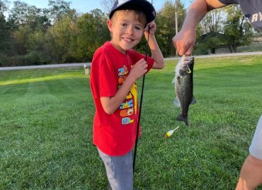 A boy in a red shirt proudly holds a fishing rod next to a caught fish.