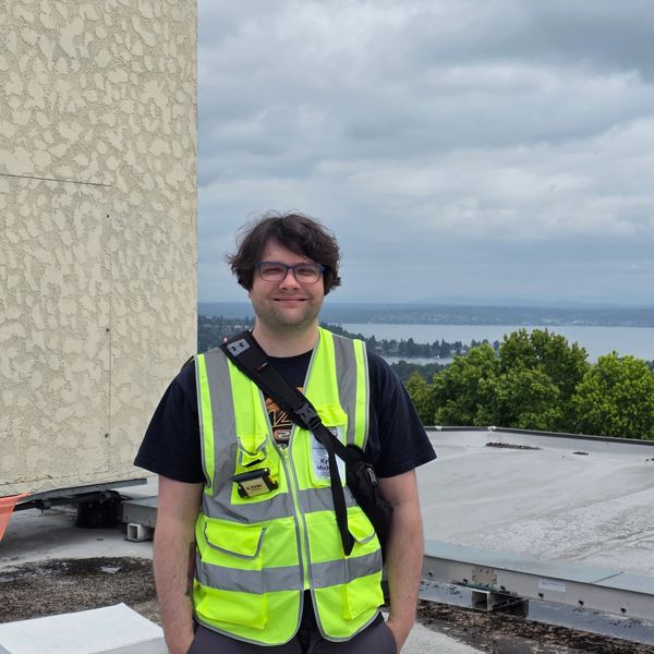 Man in safety vest standing on rooftop with trees and water in background.