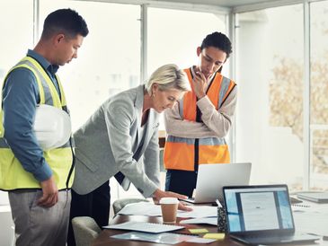 Three professionals reviewing plans in an office with laptops and documents.