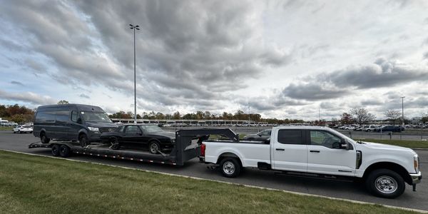 White truck towing trailer with two vehicles under cloudy sky.