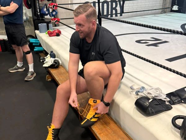 Man tying bright boxing shoes beside a boxing ring inside a gym.