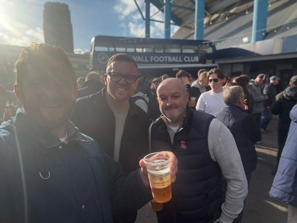 Three men enjoying drinks outside a football stadium on a sunny day.