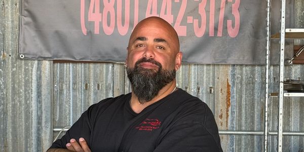 Bearded man in black shirt stands confidently with arms crossed in front of a corrugated metal wall.