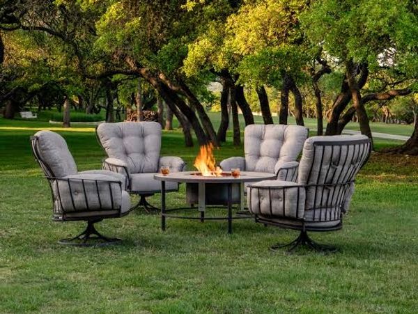 Outdoor seating area with cushioned chairs around a fire pit in a green park.
