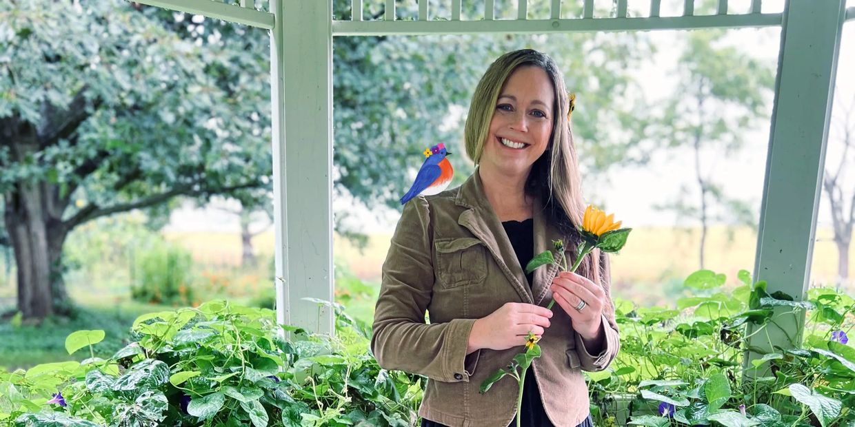 Woman smiling, holding a sunflower with a cartoon bird on her shoulder.