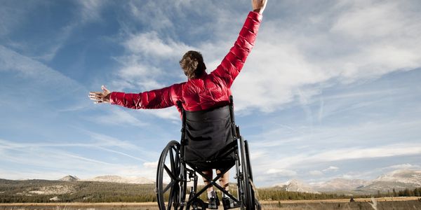 A man in a wheelchair with his back facing us and arms outstretched looking up at the blue sky