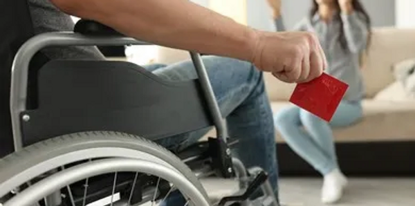 A man in a wheelchair holding a red condom wrapper with a smiling woman sitting on a couch. 