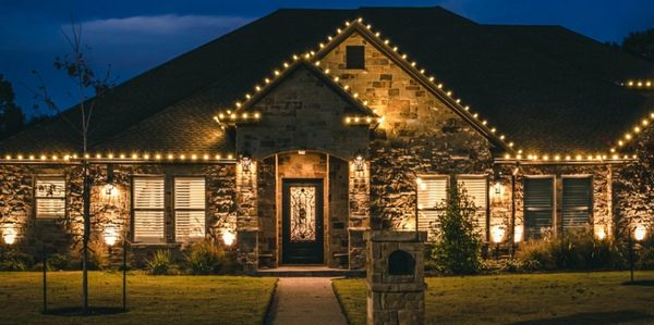 Stone house lit with warm lights and string lights on roof edges at night.