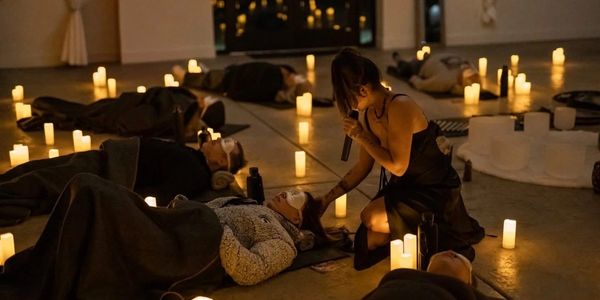 A woman sings into a microphone surrounded by candlelit people lying down.