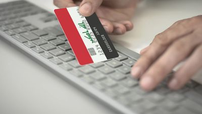 Person holding an Iraqi flag-themed credit card over a keyboard for online payment.
