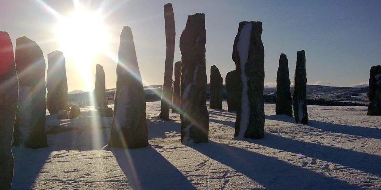 Photo of Callanish Stones in the snow, with a bright blue sky and sun poking through the Stones
