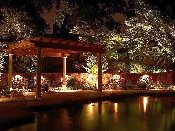 A beautifully lit garden pergola with surrounding trees and a reflective water body at night.