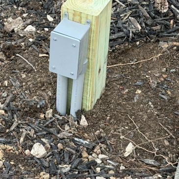 Outdoor electrical outlet box mounted on a wooden post in soil and mulch.
