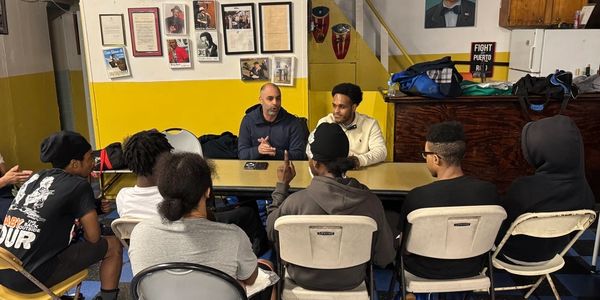 A group meeting with two men speaking to a seated audience in a room with yellow walls.