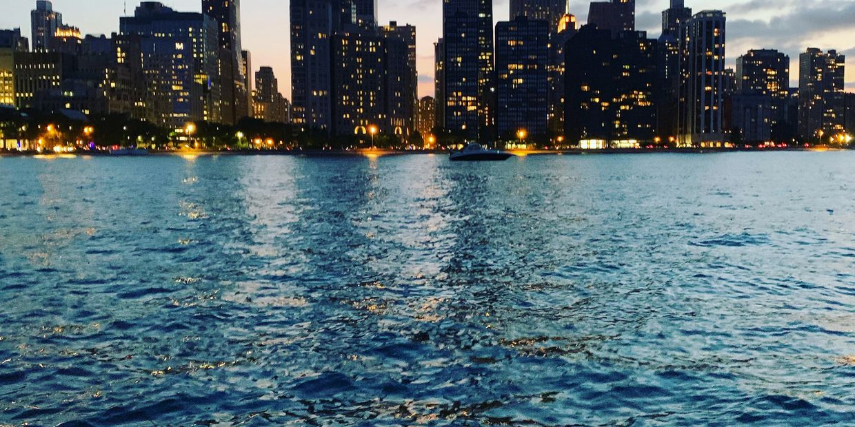 City skyline at dusk with illuminated buildings reflecting on water.