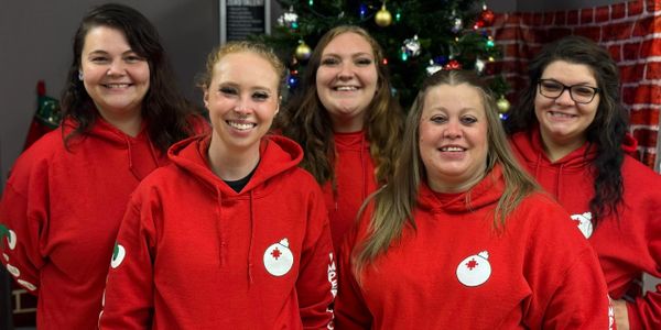 Five smiling women in red hoodies pose in front of a Christmas tree.