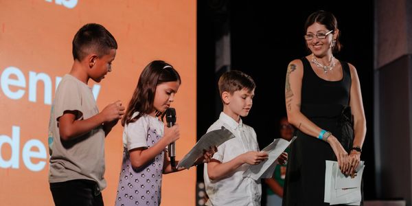Children reading from papers on stage with a smiling woman.