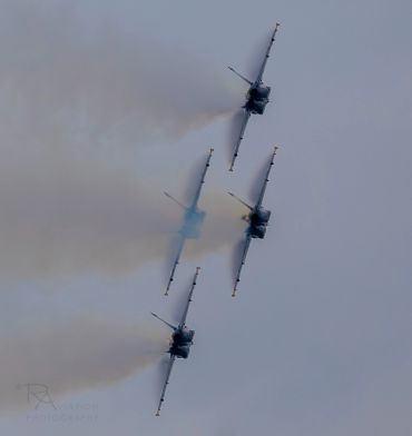 Four jets flying in tight formation with smoke trails against a gray sky.