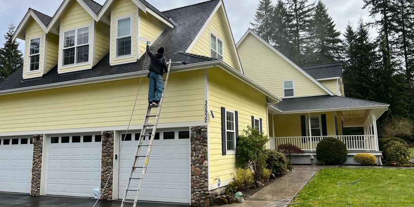 Zach soft washing a yellow house from a ladder on a rainy day.