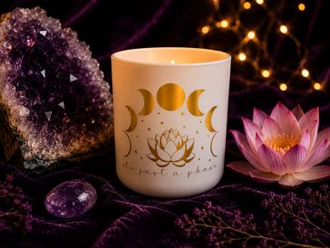 A lit candle with moon phases and lotus design beside amethyst and a pink lotus flower.