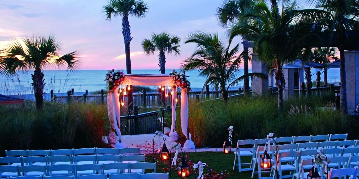 Beachside wedding setup at sunset with decorated arch and white chairs.