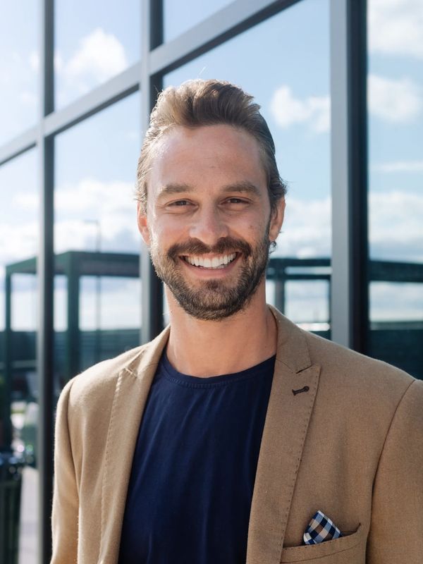 Smiling man in a tan blazer stands outdoors against glass windows.