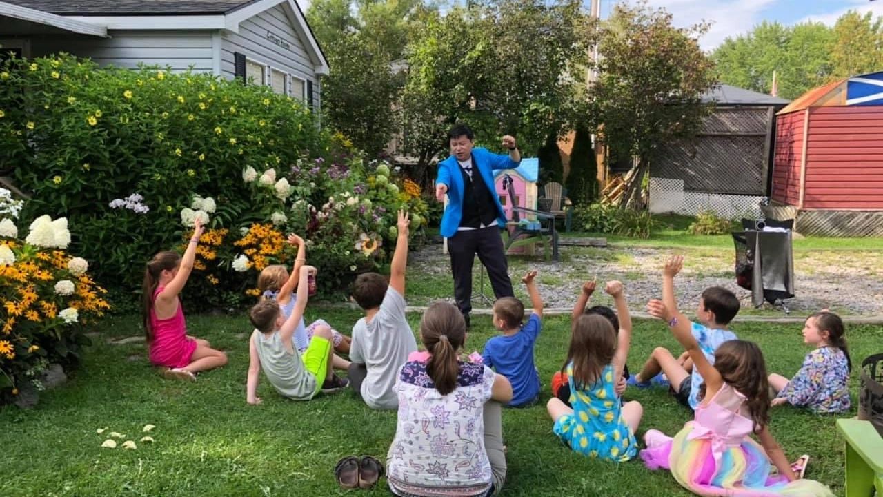 Children sitting on grass raising hands to a man in blue jacket outdoors.